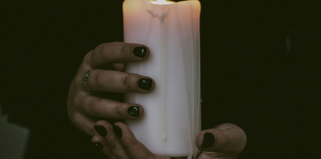 A figure in a black robe holds a lit large white cylindrical candle in hands with black fingernails.