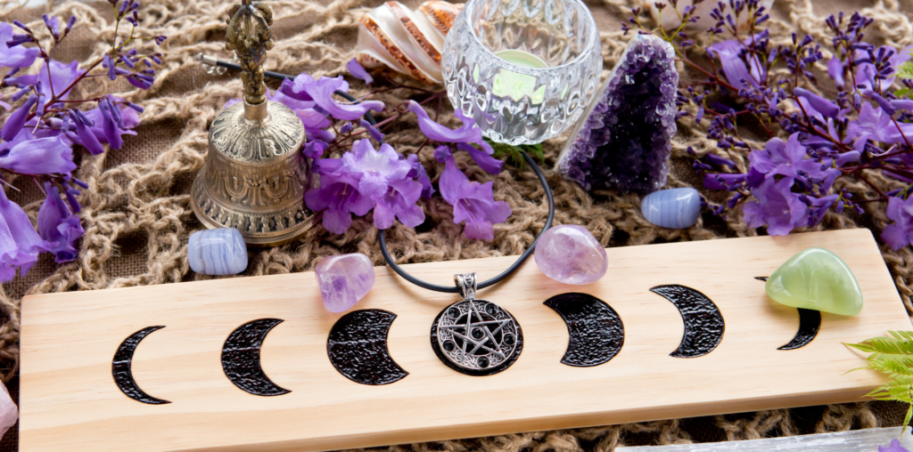 a woven jute cord pattern is on a white table. Purple flower petals are laying around a white candle, gold bell, and wooden board with black moon phase designs woodburnt onto it.